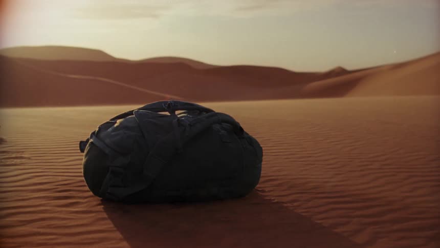 A backpack on a sandy desert with dunes the sky and clouds in the background