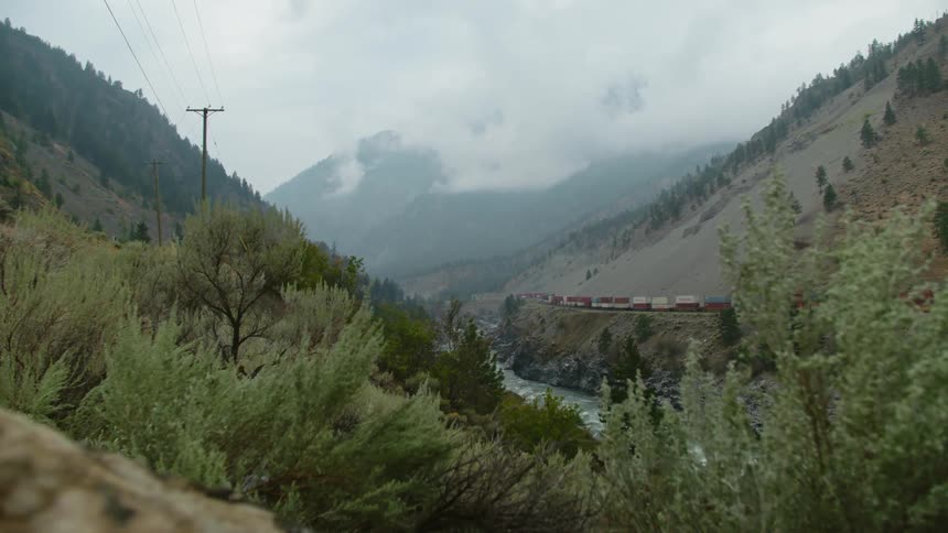A river in a valley next to a train with a mountain range the sky and clouds in the background
