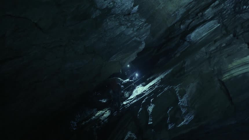 Caver rappels down steep slope in cavern lit by lights