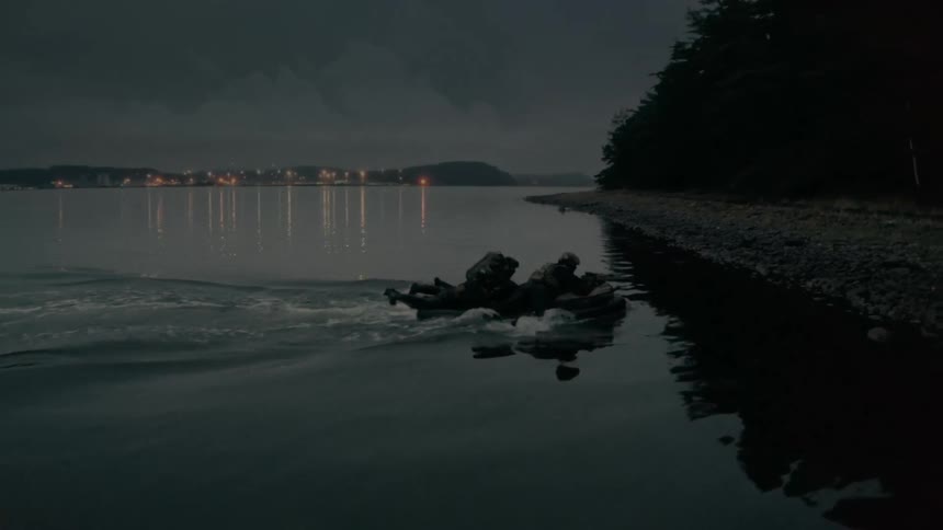 Seal team propels small raft onto pebble strewn shore near forest with fog covered town in distance