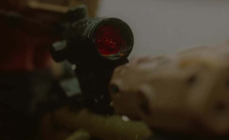 A soldier in personal protective equipment aims a firearm using a scope