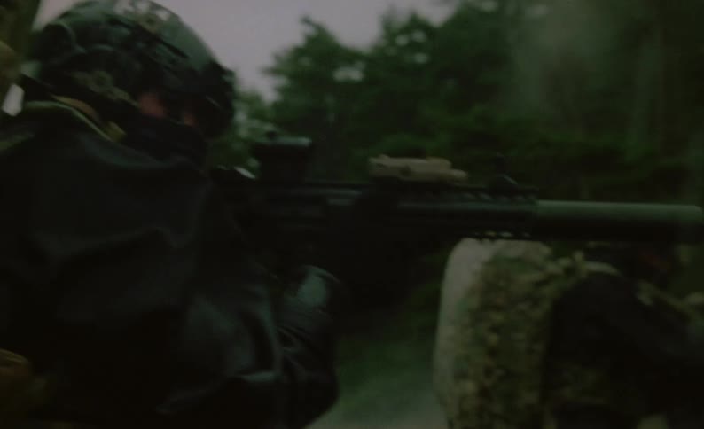 Armed and camouflaged soldiers wade out of water into woods moving in unison keeping weapons aimed ahead