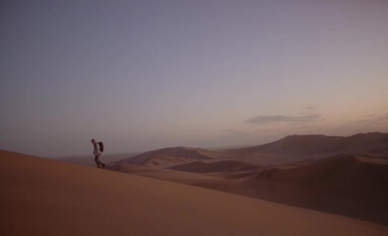 A man hikes up a sand dune in a barren desert