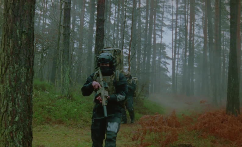 Two men in camouflage gear are carrying firearms and walking through a forest