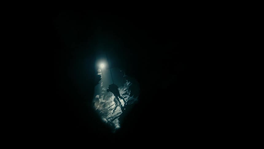 Caver hangs on rope on steep slope in lit cavern at end of dark tunnel
