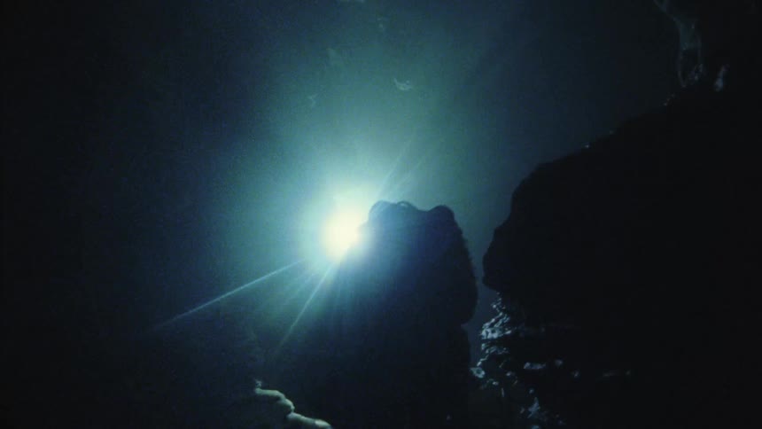 A man climbing down inside a cave using ropes is silhouetted by a large spotlight above him