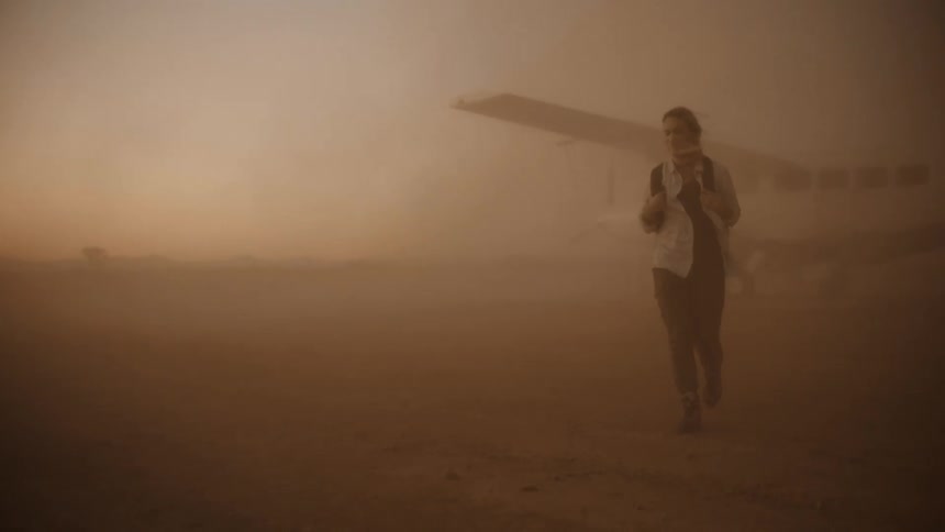 Woman wearing a white shirt and a backpack walks away from an airplane in the desert as blowing sand fills the air