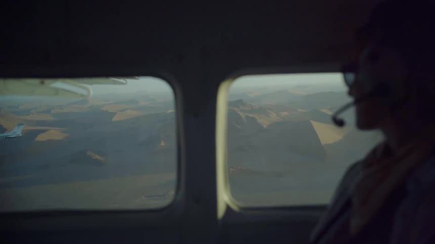 Woman wearing headset sits in plane looking at second small plane and terrain