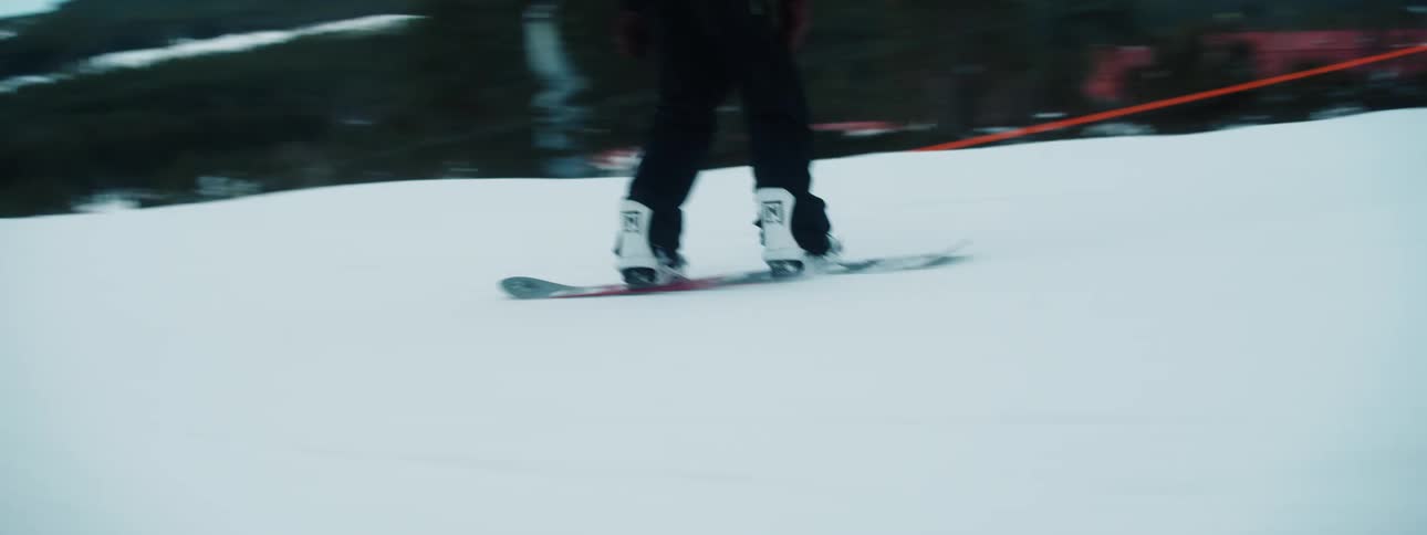 Snowboarder rides over snowboarding course passing resort with mountains and forest in background