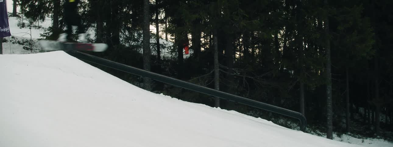 Person skiboarding over a railing on a snow covered hill near a forest at sunset