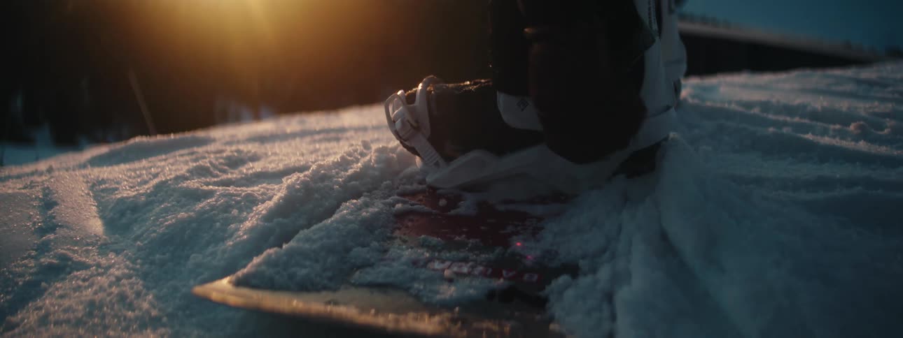 A person on a snowboard does up their boots as another person skis past them