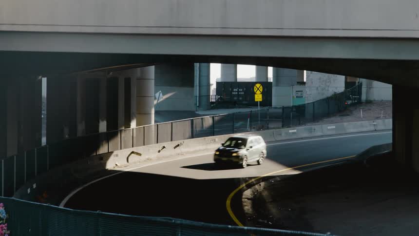 A vehicle driving on a road underneath an overpass with a sign and a train in the background