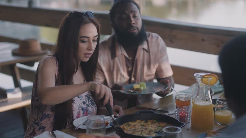 Woman with brown long hair puts food on plate for man with pink shirt and beard in restaurant next to lake