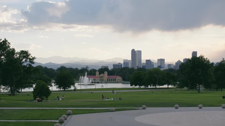 A water fountain is shooting water behind a city park where people are relaxing and picnicking