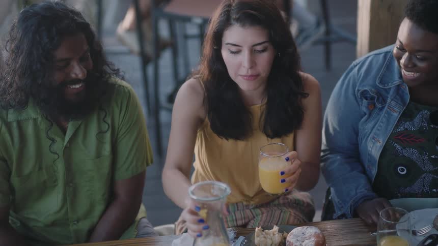 A woman sitting at a table outdoors talks with her friends while pouring a glass of orange juice