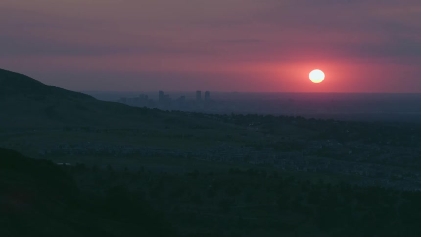 Bright sun rises from horizon into red sky surrounded by purple with fog covered city in background and silhouetted slope
