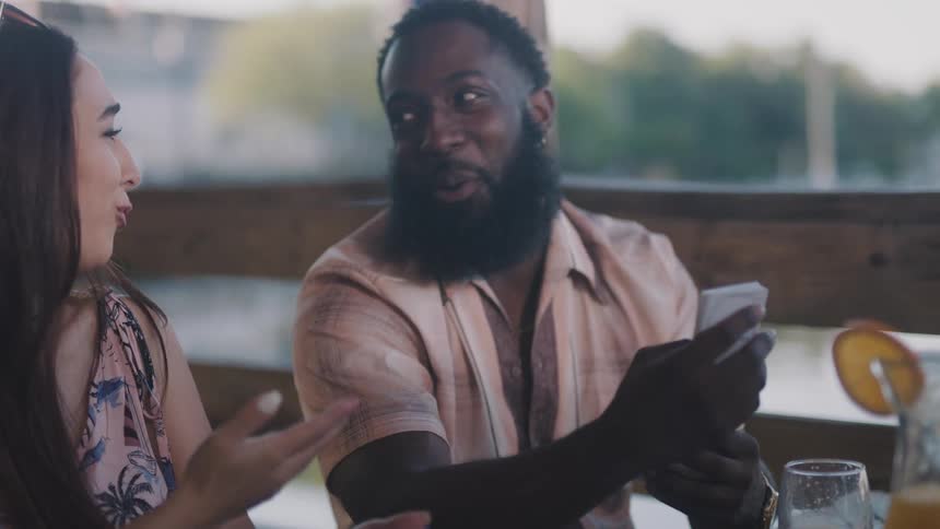 Man with beard and pink shirt sits at restaurant table holding cards and talking woman with long straight hair laughing