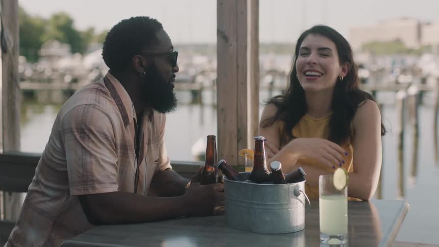 Man with beard sunglasses and pink shirt sits at table holding beer bottle and talking to woman wearing yellow blouse in restaurant next to lake