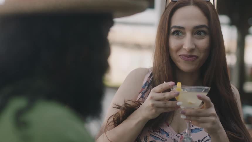 A woman talking to a man with a hat with sunglasses on her head and drinking a drink in a glass