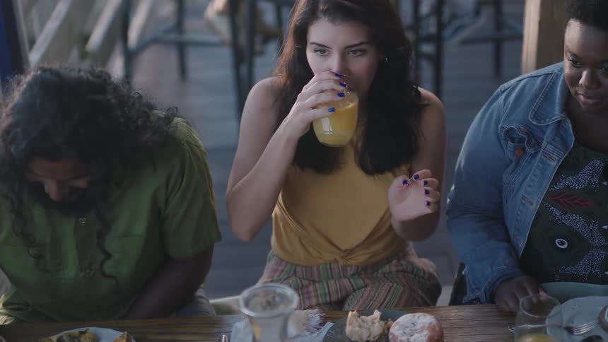 Three people are sitting at a table eating and the woman in the middle drinks her orange juice