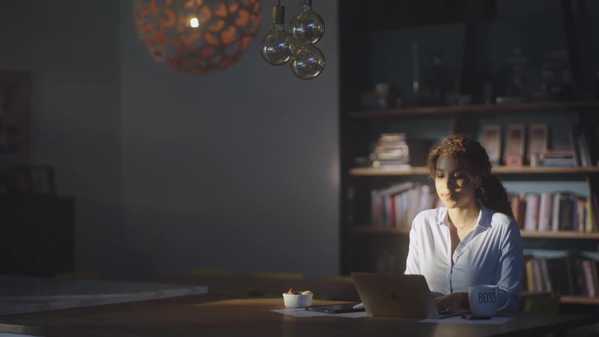 Woman with curly hair and shirt sits at table using laptop in front of bookshelf