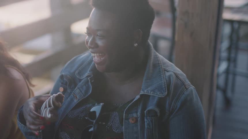 A woman wearing a denim jacket and holding a donut is smiling and laughing