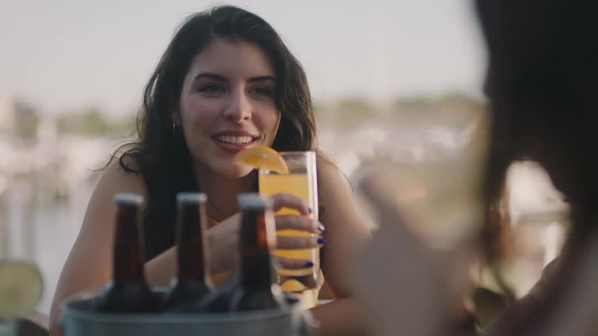 Woman with brown wavy hair drinks orange juice sitting at table with friends drinking beer at cafeteria next to lake