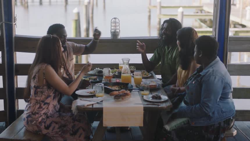 A group of friends are talking and eating at a restaurant table overlooking a lake