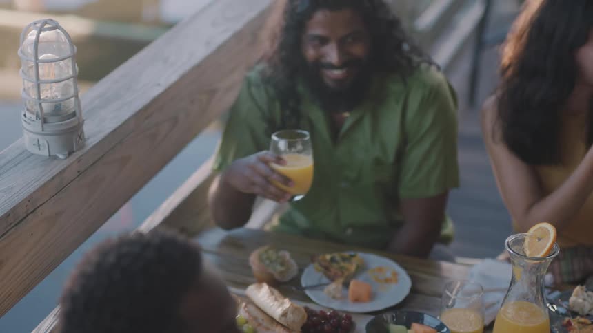 A group of people drink orange juice while eating at a wooden table outdoors during the day