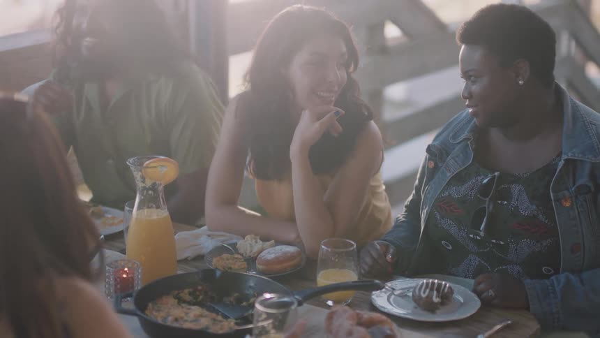 Friends sit outdoors near wooden rail laughing and talking over brunch