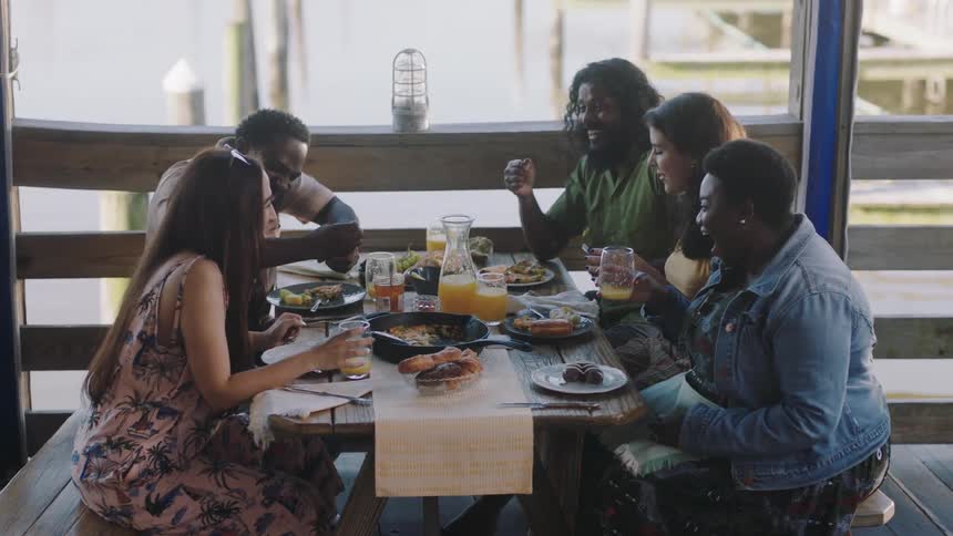 Five people are sitting at a table with plates of food and drinking orange juice and talking