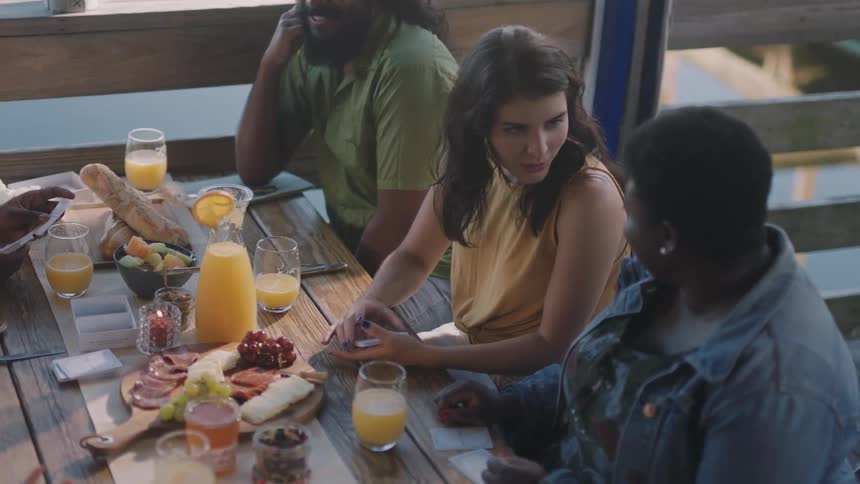 Friends sit at table spread with meat and cheese tray smiling and talking with water showing between wooden slats of rail