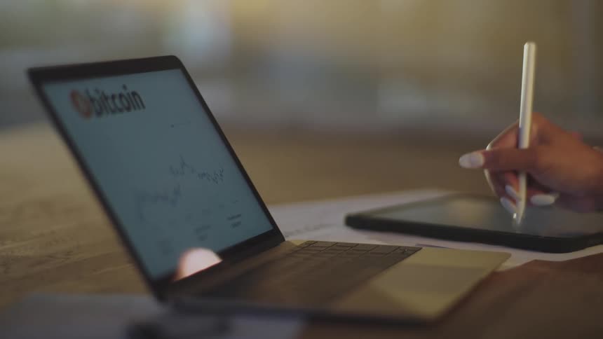 Woman works at desk making notes on tablet with bitcoin website on the laptop screen