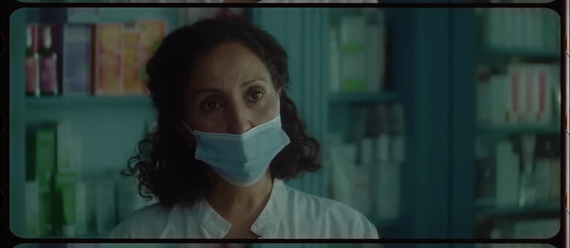 A female clerk wearing a mask in a small town drug store interacts with an unseen customer and then stares blankly