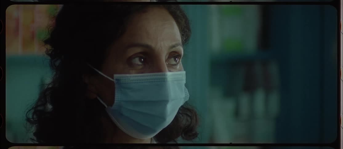 A woman with dark hair is wearing a surgical mask and moving her eyes in a concerned manner