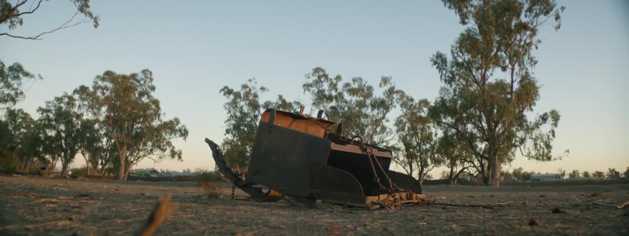 Broken part of metal trailer sits on ground under clear sky near trees