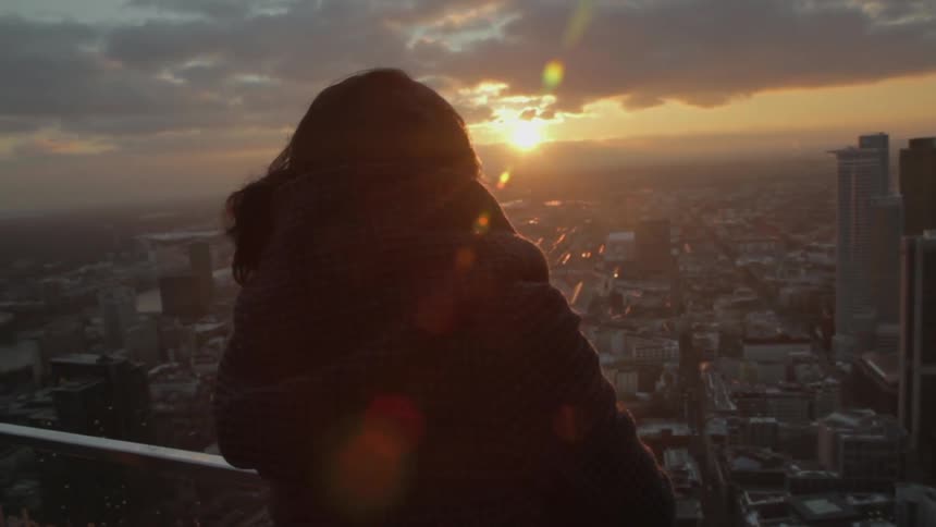 A woman stands on the balcony overlooking a city at sunset