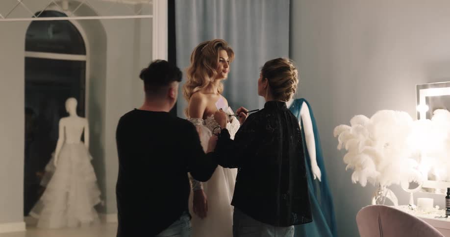 A woman stands in a design studio wearing a wedding gown as a woman applies makeup and a designer adjusts her gown