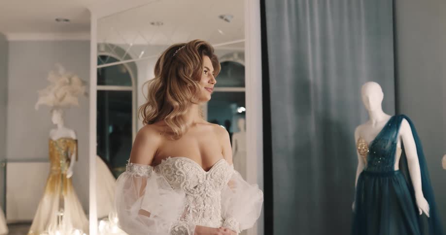 Woman in wedding dress and with brown wavy hair stands as shop assistant puts on makeup on her in bridal shop
