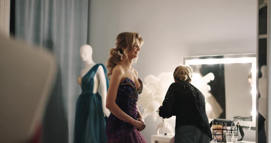 A woman applies makeup to a model in an evening gown