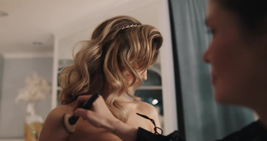 A woman in a room wearing a wedding dress with another woman applying makeup to her body with a brush with a wall in the background