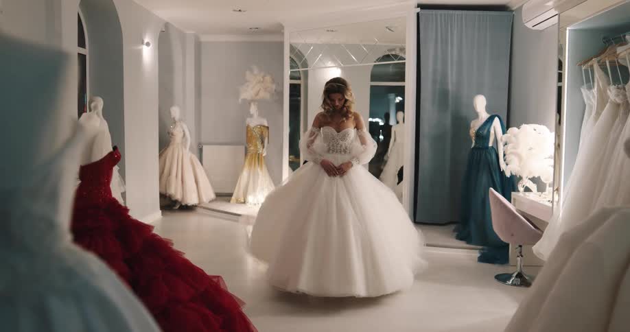 Woman with brown wavy hair tries on wedding dress in fitting room and looks in mirror talking to male shop assistant with black shirt