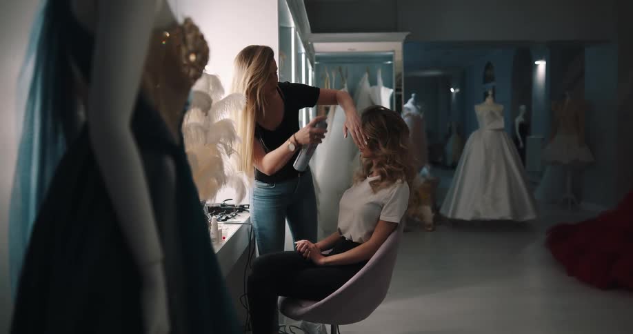 A woman in a room doing another womans hair with a spray can with mannequins and a wall in the background