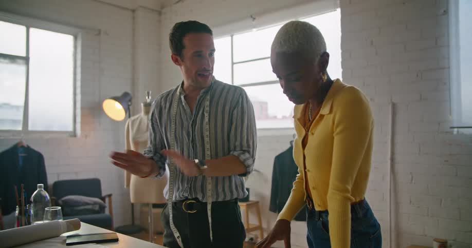 A black woman wearing a yellow sweater is talking to a man with a tape measure around his neck looking at a table