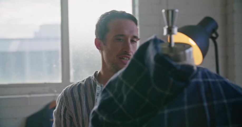 A man with a measuring tape in a room with a wall a window and a lamp in the background
