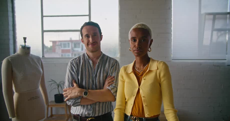 Male and female designer standing next to mannequin look at camera smiling in dressmaker workshop