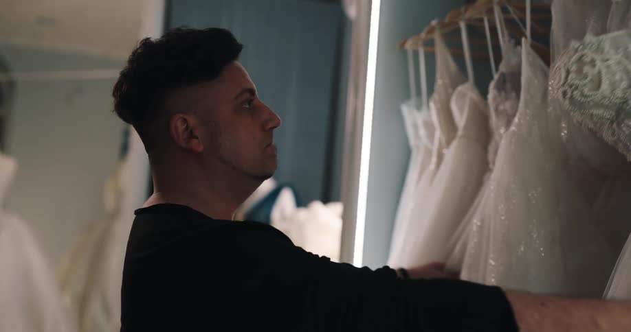 Man examines line of wedding dresses on hangers before choosing one an carrying it from room