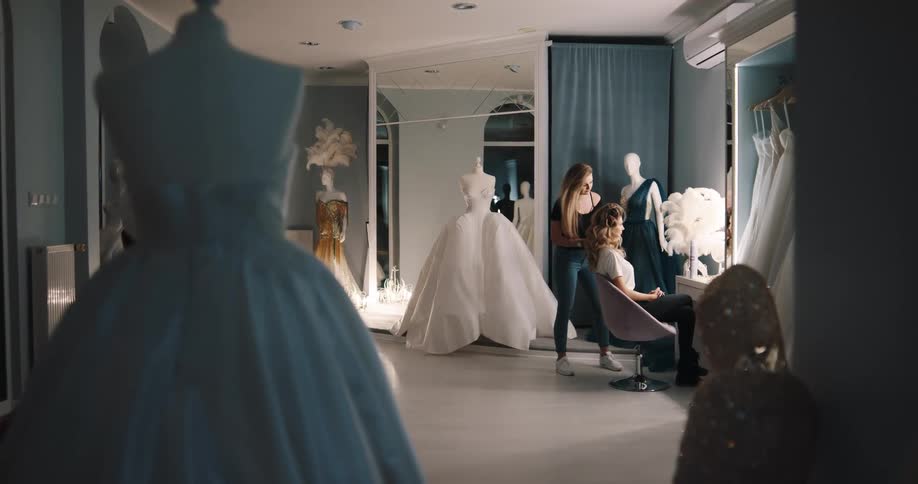 A woman in a room with a mirror doing another womans hair thats sitting on a chair with a wall in the background