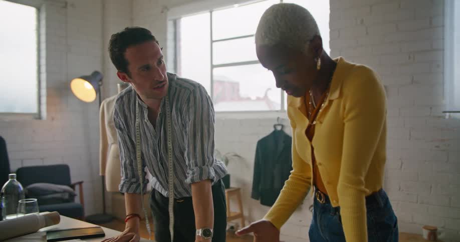 A black woman wearing a yellow sweater is talking to a man with a tape measure around his neck looking at papers on a table