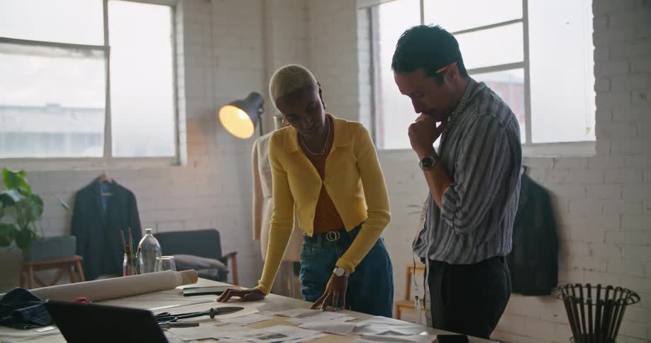 A woman wearing a yellow sweater is talking to a man with a tape measure around his neck and looking at papers on a table